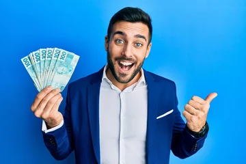 Fotobehang Young hispanic businessman wearing business suit holding brazilian real banknotes pointing thumb up to the side smiling happy with open mouth © Krakenimages.com