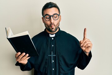Young hispanic priest man holding bible with finger up making fish face with mouth and squinting...