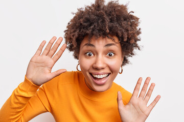 Expressive playful curly African American woman smiles broadly raises palms high foolishes around...