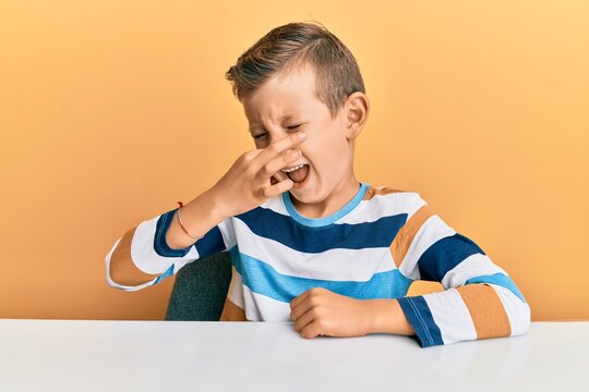 Adorable caucasian kid wearing casual clothes sitting on the table smelling something stinky and disgusting, intolerable smell, holding breath with fingers on nose. bad smell