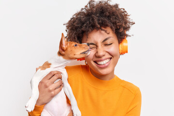 Nice dog licks face of female owner with tenderness expresses love. Happy curly haired woman spends time together with favorite pet listens music in wireless headphones isolated on white wall - obrazy, fototapety, plakaty