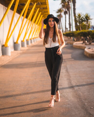 Lifestyle, a young brunette walking through the Mediterranean in heels, pants and black hat on a yellow promenade