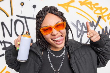Cool dark skinned woman sticks out tongue makes yo gesture sprays paint from aerosol bottle creats...