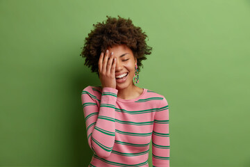 Fototapeta na wymiar Studio shot of happy African American woman makes face palm smiles broadly has fun laughs out loudly at something very funny dressed in casual striped jumper isolated over bright green background