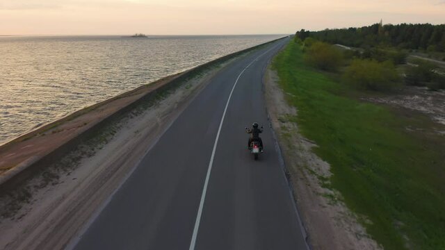 A girl in motorcycle clothes rides cruiser motorbike along the road along the sea at sunset. Aerial view
