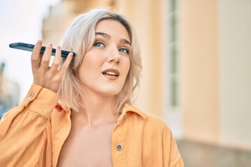 Young blonde girl smiling happy listening audio message using smartphone at the city.