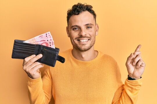 Young hispanic man holding wallet with yuan chinese banknotes smiling happy pointing with hand and finger to the side
