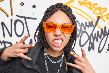 Funky ethnic teenage girl clenches teeth makes yo gesture feels cool has dreadlocks hairstyle wears orange sunglasses black jacket and metal chains around neck stands against graffiti background - obrazy, fototapety, plakaty