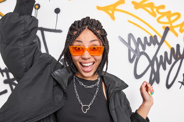 Cheerful hipster girl dances carefree keeps arms raised smiles broadly wears orange sunglasses black jacket poses against graffiti wall expresses positive emotions. Youth subculture and lifestyle - obrazy, fototapety, plakaty