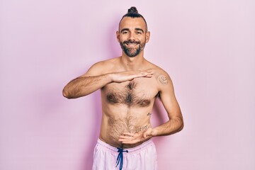Fototapeta premium Young hispanic man shirtless wearing swimsuit gesturing with hands showing big and large size sign, measure symbol. smiling looking at the camera. measuring concept.
