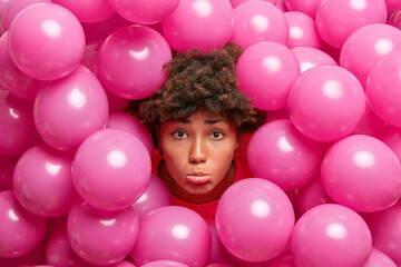 Fototapeta na wymiar Displeased curly haired young woman sticks out head through inflated balloons looks sadly at camera expresses negative emotions being in bad mood. Party decorations concept. Spoiled holiday.
