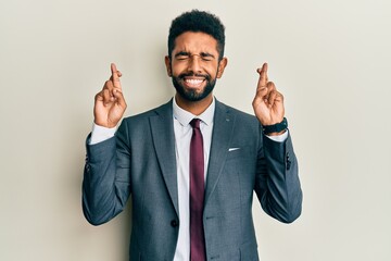 Handsome hispanic man with beard wearing business suit and tie gesturing finger crossed smiling with hope and eyes closed. luck and superstitious concept.