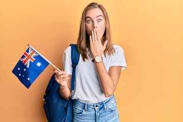 Beautiful blonde woman exchange student holding australia flag covering mouth with hand, shocked...