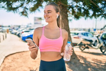 Young blonde sportswoman using smartphone and drinking bottle of water at the city.