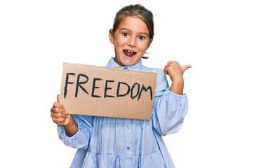 Little beautiful girl holding freedom banner pointing thumb up to the side smiling happy with open mouth