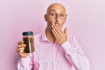 Bald man with beard holding soluble coffee covering mouth with hand, shocked and afraid for...
