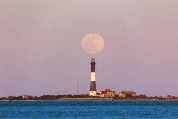 Fototapeta premium Full moon rising through the Belt of Venus behind a tall stone lighthouse at sunset. Fire Island, Long Island New York