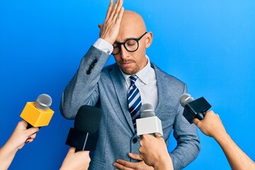 Bald man with beard being interviewed by reporters holding microphones surprised with hand on head...