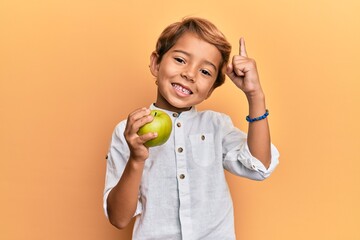 Adorable latin kid holding green apple smiling with an idea or question pointing finger with happy...