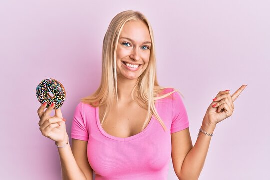 Young blonde girl holding donut smiling happy pointing with hand and finger to the side