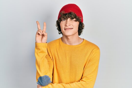 Handsome young man wearing wool hat smiling with happy face winking at the camera doing victory sign. number two.