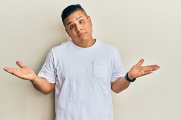 Young latin man wearing casual white t shirt clueless and confused expression with arms and hands...