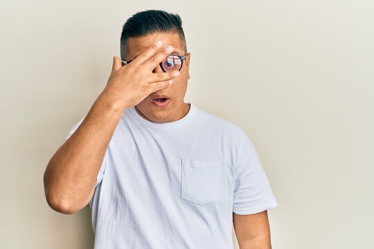 Young latin man wearing casual clothes and glasses peeking in shock covering face and eyes with hand, looking through fingers afraid