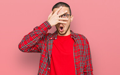 Hispanic young man wearing casual clothes peeking in shock covering face and eyes with hand,...