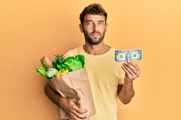 Handsome man with beard holding groceries and 1 american dollar banknote relaxed with serious expression on face. simple and natural looking at the camera.