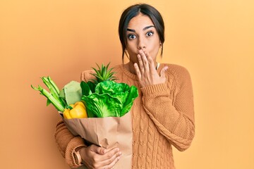 Young brunette woman holding paper bag with bread and groceries covering mouth with hand, shocked...