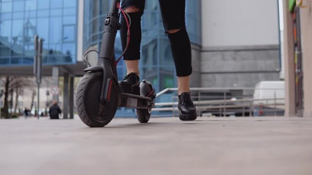Young stylish blonde girl in a beige trench coat and jeans gets her foot on an electric scooter and rides with a modern high-rise on the background. Kind of feet, shoes and wheels