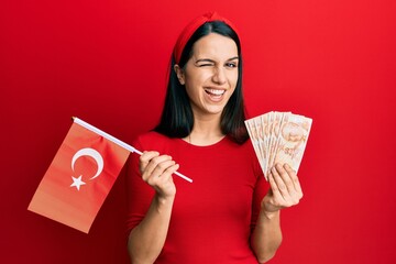 Young hispanic woman holding turkey flag and liras banknotes winking looking at the camera with...
