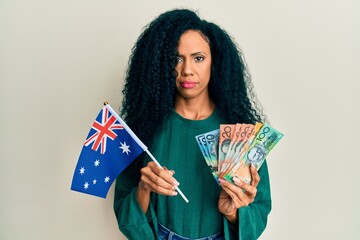 Middle age african american woman holding australian flag and dollars relaxed with serious expression on face. simple and natural looking at the camera.