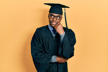 Young african american man wearing graduation cap and ceremony robe with hand on chin thinking...