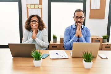 Middle age hispanic woman and man sitting with laptop at the office praying with hands together asking for forgiveness smiling confident.