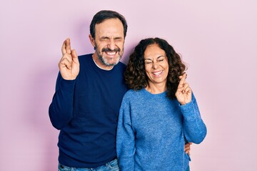 Fototapeta premium Middle age couple of hispanic woman and man hugging and standing together gesturing finger crossed smiling with hope and eyes closed. luck and superstitious concept.