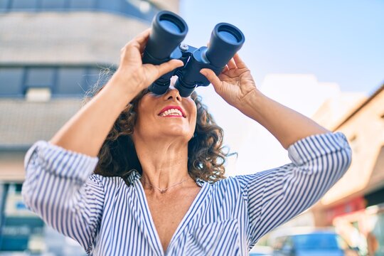 Middle age hispanic woman smiling happy looking for new opportunity using binoculars at the city.