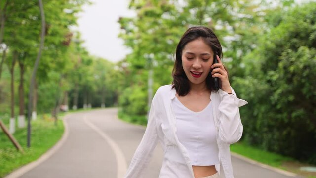 Pretty young asian woman walking on the road in the park outdoor talking on the mobile phone happy people using phone in the nature relax lifestyle 