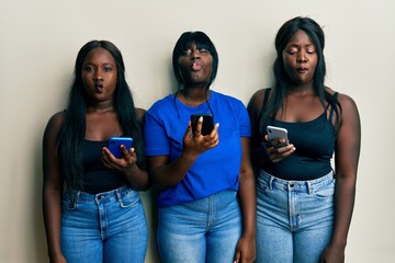 Three young african american friends using smartphone making fish face with lips, crazy and comical gesture. funny expression.