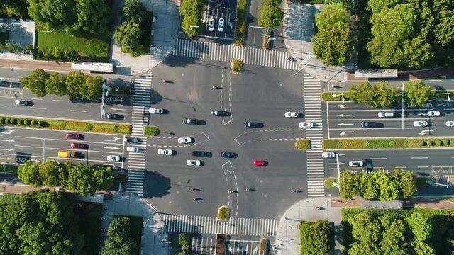 Aerial top down view of an intersection crossroads with traffic, moving cars of different colors and ebikes, surrounded by green trees. Camera moves down