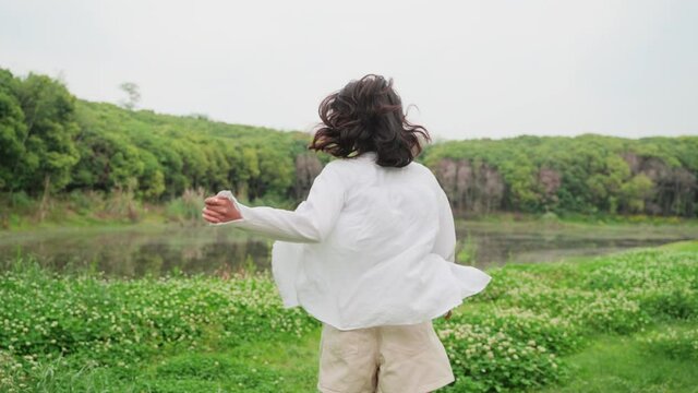Charming young asian girl in white shirt running in the spring nature field by the lake turn back looking at camera smile lovely young woman enjoy the nature freedom lifestyle 
