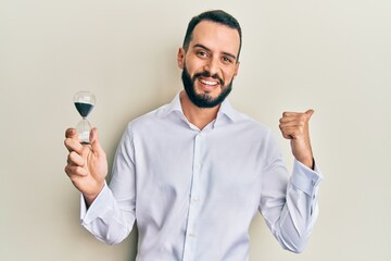 Young man with beard holding sand clock pointing thumb up to the side smiling happy with open mouth