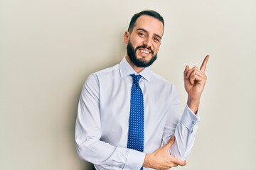 Young man with beard wearing business tie with a big smile on face, pointing with hand and finger to the side looking at the camera.