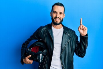 Naklejka premium Young man with beard holding motorcycle helmet surprised with an idea or question pointing finger with happy face, number one