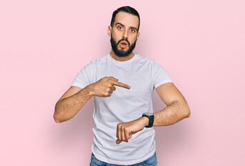 Young man with beard wearing casual white t shirt in hurry pointing to watch time, impatience,...