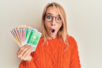 Beautiful blonde woman holding norwegian krone banknotes scared and amazed with open mouth for surprise, disbelief face