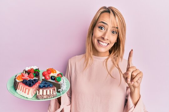 Beautiful blonde woman holding cake slices smiling with an idea or question pointing finger with happy face, number one