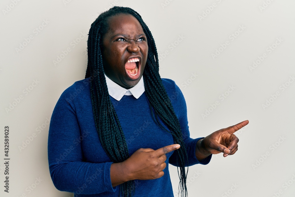 Sticker Young black woman with braids pointing up with fingers to the side angry and mad screaming frustrated and furious, shouting with anger. rage and aggressive concept.