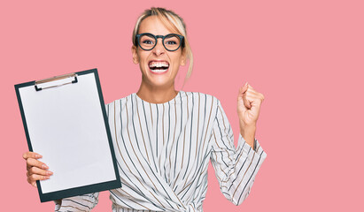 Beautiful blonde business woman holding clipboard with blank space screaming proud, celebrating victory and success very excited with raised arms