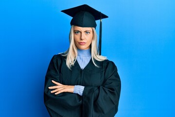 Beautiful blonde woman wearing graduation cap and ceremony robe skeptic and nervous, disapproving expression on face with crossed arms. negative person.